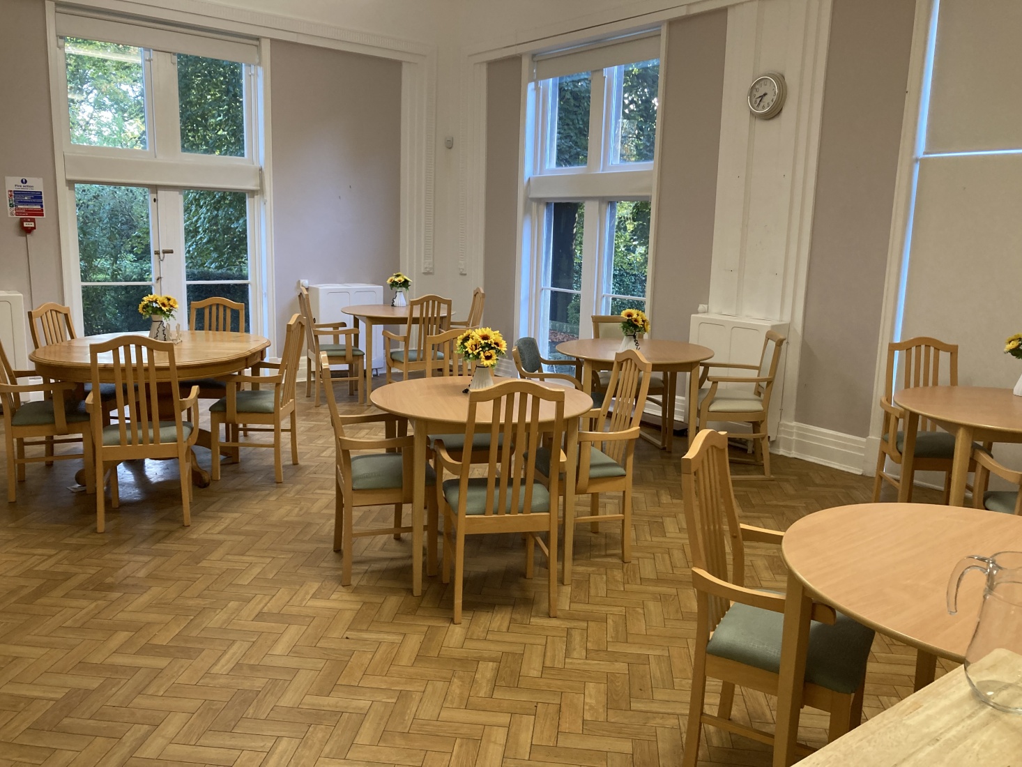 A dining room with wooden floor with dining chairs around round tables. There are tall windows with roller blinds.