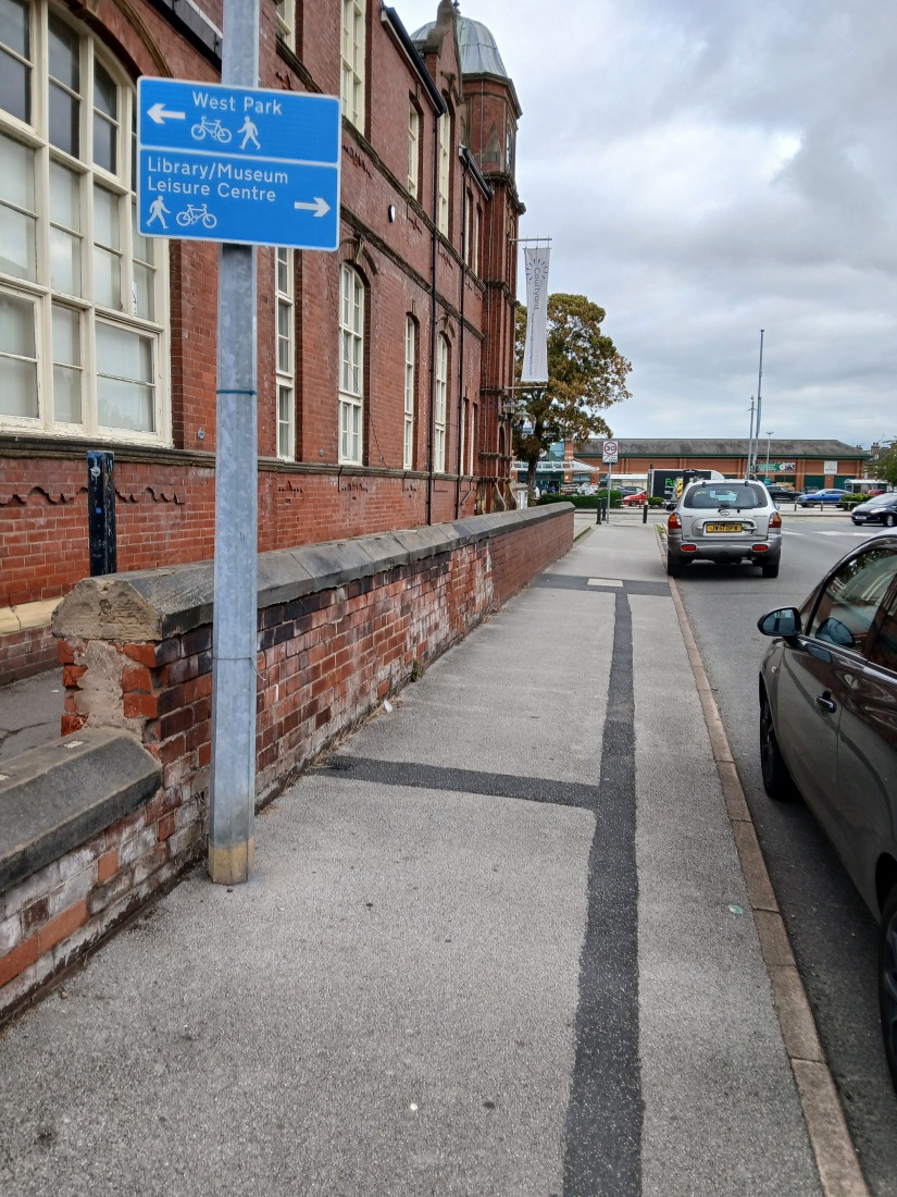 The side of a brick building with a public footpath towards the entrance. There are a few cars parked on the road.