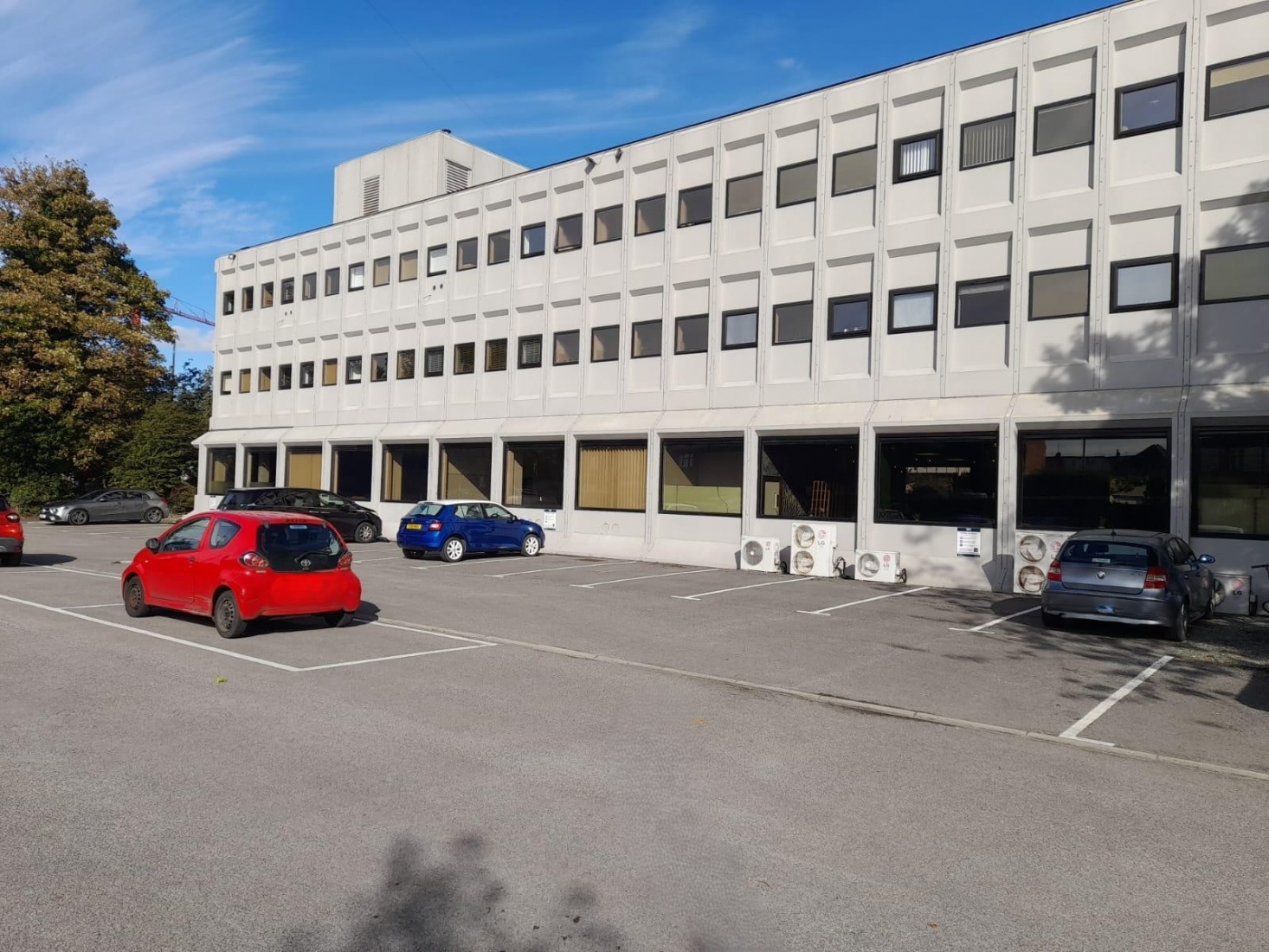 A car park in front of a pale geometric 3 story building with lots of windows
